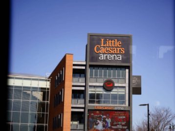 The Little Caesars arena sign, shot from inside a bus, with clear blue sky weather conditions.